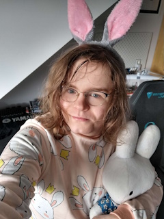 A photo of isocosa wearing a sweater with bunnies on it and a pair of bunny ears. She's also holding a Nijntje/Miffy plushie, who is also a bunny.