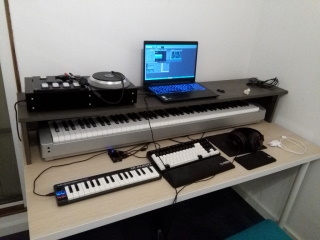 A desk with a shelf laying on top of it. On the desk itself there's a small 32-key MIDI keyboard, a regular computer keyboard, a mouse and a phone charger. On top of the shelf, there's a IIDX controller, a laptop, a keycap remover, a bunny figurine and a bunch of cables. Under the shelf there's a large full-sized MIDI keyboard.