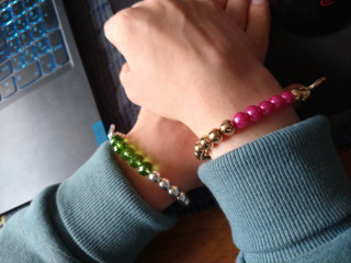 A photo of arms with a bracelet on each. The left bracelet has silver beads, and a section with green gem-like beads. The right bracelet has golden beads, and a section with pink beads.