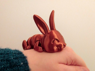 A photo of a 3D-printed articulated rabbit toy in the color of brass. The toy is laying on the top of isocosa's hand, as if it's sleeping.
